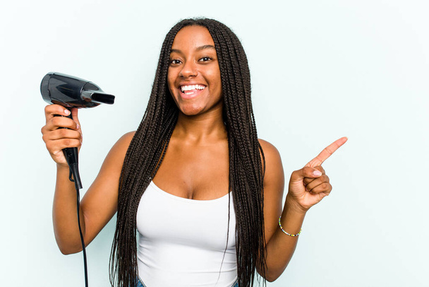 Young African American woman holding a hairdryer isolated on blue background smiling and pointing aside, showing something at blank space. - Foto, imagen