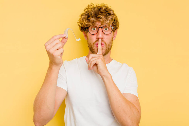 Young caucasian man wearing hearing aid isolated on yellow background keeping a secret or asking for silence. - Photo, Image