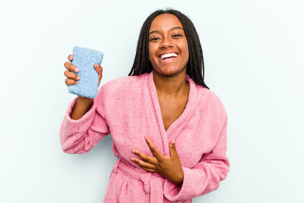 Молодая афроамериканка, держащая губку на синем фоне, громко смеется, держа руку на груди.. - Фото, изображение