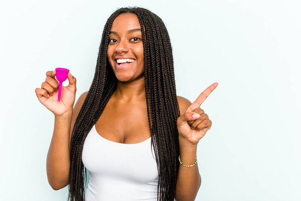 Młoda Afroamerykanka trzymająca kubek menstruacyjny odizolowana na niebieskim tle uśmiechnięta i skierowana na bok, pokazująca coś w pustej przestrzeni. - Zdjęcie, obraz