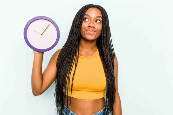 Młoda Afroamerykanka trzymająca zegar na niebieskim tle marząca o osiągnięciu celów - Zdjęcie, obraz