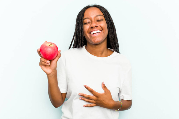 Junge Afroamerikanerin, die einen Apfel auf blauem Hintergrund hält, lacht laut und hält die Hand auf der Brust. - Foto, Bild