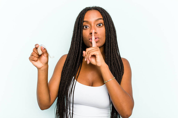 Elinde işitme cihazı tutan genç Afrikalı bir kadın mavi arka planda bir sır saklıyor ya da sessizlik istiyor.. - Fotoğraf, Görsel