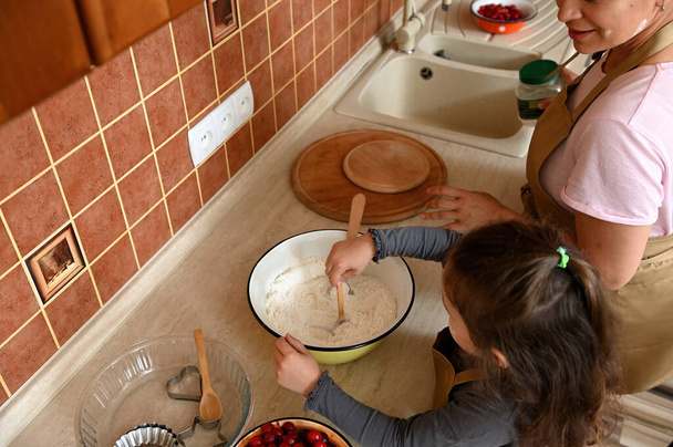 Вид сверху: очаровательная маленькая девочка, стоящая у столешницы, смешивающая тесто в поисках вкусного праздничного вишневого пирога, помогающая маме на кухне. Ребенок учится кулинарии. Мама и дочь готовят вместе - Фото, изображение