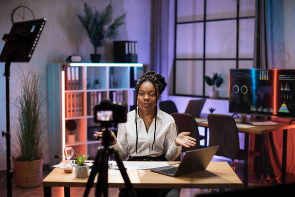 Cheerful confident african american businesswoman, broker in white shirt sitting in front of camera in evening office during recording video for business vlog. - Foto, afbeelding