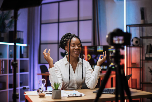 Relaxed motivated african american businesswoman in formal wear meditating with closed eyes and mudra hands in front of camera during recording video for business vlog in evening office - Fotoğraf, Görsel