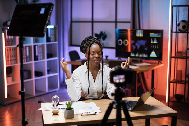 Relaxed motivated african american businesswoman in formal wear meditating with closed eyes and mudra hands in front of camera during recording video for business vlog in evening office - 写真・画像