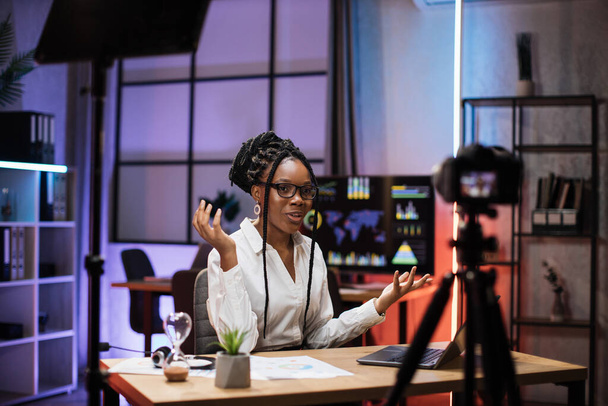 Confident african american businesswoman, broker in white shirt sitting in front of camera in evening office during recording video for business vlog. - Foto, Bild
