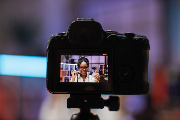 Focus on camera screen with attractive confident experienced smart female african american office manager in headset recording video vlog for internet audience in evening office showing thumb up. - Fotoğraf, Görsel