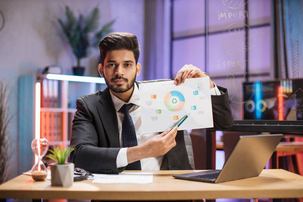 Confident arab financial expert office worker in suit working on laptop, showing paper report with graphs and charts during video meeting with colleagues in office. - Photo, Image