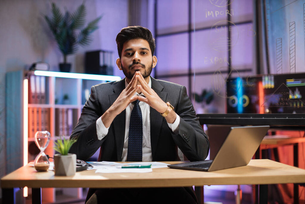 Indian bearded man in formal suit sitting at desk with modern laptop and looking at camera with serious facial expression. Concept of people and office work. - 写真・画像