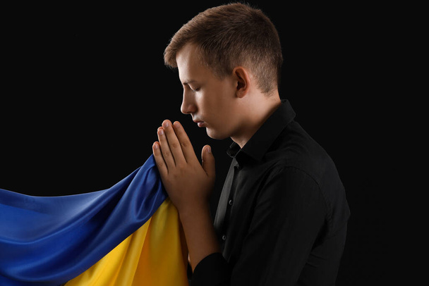 Junger Mann mit Fahne der Ukraine betet auf schwarzem Hintergrund - Foto, Bild