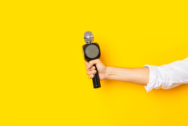 La mano del hombre sostiene un micrófono en un agujero en un fondo de cartón amarillo brillante desgarrado. - Foto, imagen