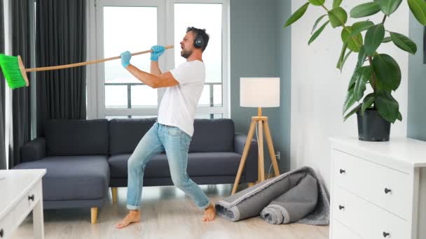 Hombre caucásico en auriculares limpiando la casa y divirtiéndose bailando y cantando con una escoba. Movimiento lento - Imágenes, Vídeo