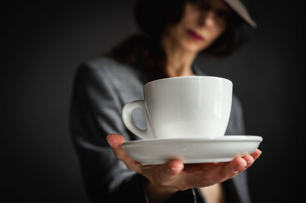 dziewczyna w kapeluszu i szarej kurtce trzyma biały kubek z poranną kawą i herbatą w rękach, na szarym tle. Skupienie selektywne - Zdjęcie, obraz