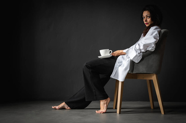 Fiatal, csinos, érzelmes lány ül egy modern székben, élvezi a kávét, a teát szürke háttérrel, oldalnézetet, figyelmes, gondolkodó. Írástér - Fotó, kép