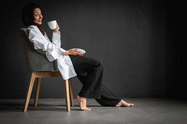 Young pretty, emotional girl sits in a modern chair, enjoys coffee, tea on a gray background, side view, thoughtful, thinking. Space for writing - Foto, immagini