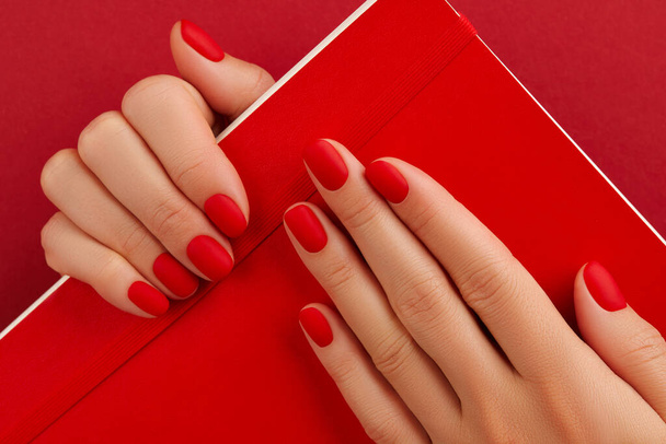 Female hand with manicure holding notepad. Red matt nail design. Beauty treatment body care concept - Photo, Image