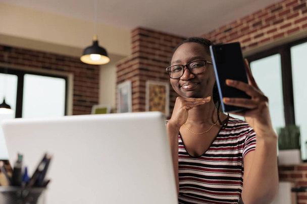 Fiatal nő arcszelfit készít okostelefonon, pózol, mosolyog a kamera előtt. Távoli diák portréfotózás a mobiltelefon beltéri, távoli munkavállaló, amelynek video chat - Fotó, kép