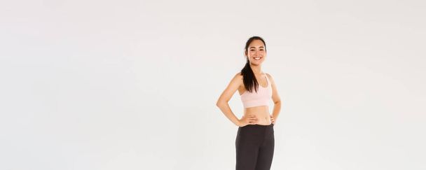 Full length of sweaty smiling fitness girl, female athelte in active wear looking pleased while workout, training in gym, satisfied with exercises, coach helping gain perfect body, white background. - Photo, image