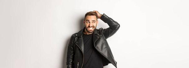 Confused macho guy in black leather jacket looking awkward and unsure, scratching head puzzled and stare left, white background. - Photo, Image