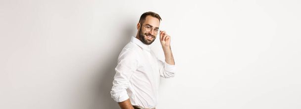 Handsome businessman turn at camera and looking confident, smiling cheeky, standing over white background. - Foto, imagen
