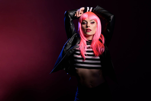 Sexy beautiful adult with pink hair wearing leather jacket in studio with dark light, posing over background. Confident woman with stylish rocker clothes and perfect makeup look, fashion style. - Photo, image