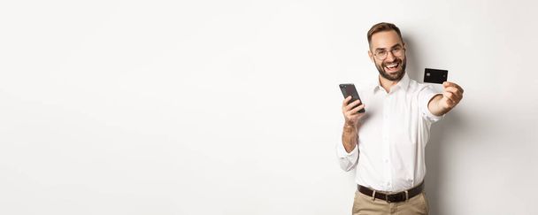 Pago en línea y de negocios. Hombre emocionado mostrando su tarjeta de crédito mientras sostiene el teléfono inteligente, de pie satisfecho contra el fondo blanco. - Foto, imagen