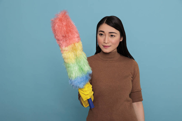 optimista feliz asiático ama de casa en amarillo guantes de goma celebración polvo cepillo aislado en un fondo azul, limpieza casa concepto, sensación alegre feliz y positivo - Foto, Imagen