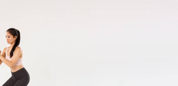 Повнометражний вид збоку зосередженої тонкої азіатської дівчини, що займається фітнесом, жіноча атлетична застібка рук разом і виконувати присідання вправи з розтягнутою смугою опору, тренувальне обладнання
. - Фото, зображення