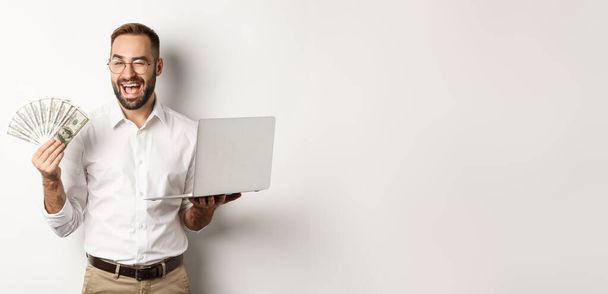 Бизнес и электронная коммерция. Уверенный бизнесмен показывает, как работают в Интернете, подмигивая, держа деньги и ноутбук, стоя на белом фоне. - Фото, изображение