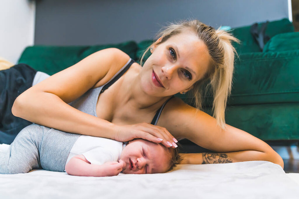 Mujer caucásica con el pelo rubio descansando en el suelo en su lado acariciando a su bebé recién nacido mirando a la cámara sonriendo. Pround madre cariñosa. Tiro interior horizontal. Foto de alta calidad - Foto, imagen
