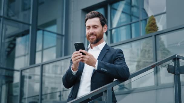 Uśmiechnięty mężczyzna stojący na zewnątrz trzymający telefon komórkowy przeglądający portale społecznościowe szczęśliwy zamyślony biznesmen myśli, że sny piszą e-mail przez telefon na czacie z klientem lub przyjacielem za pomocą smartfona - Materiał filmowy, wideo