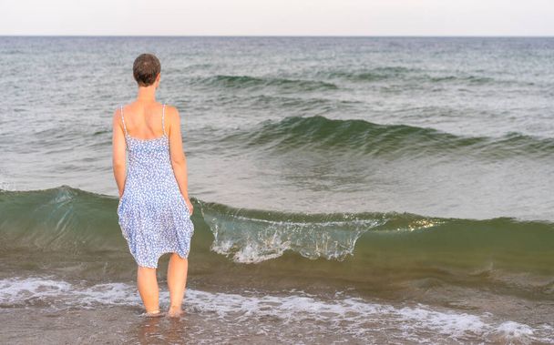 Attraente donna dai capelli corti sulla trentina indossa un abito estivo leggero cammina a piedi nudi sulla spiaggia con il mare sullo sfondo. Concetto di vacanza, svago, relax, salute fisica e mentale - Foto, immagini