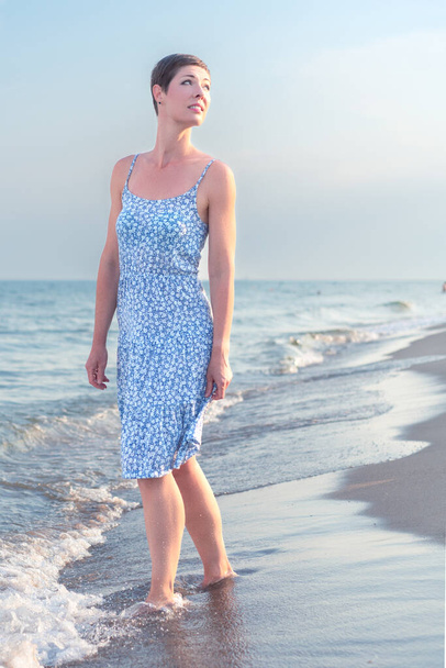 明るい夏のドレスを着た30代の魅力的な短い髪の女性は、背景に海とビーチで裸足で歩く。休暇、レジャー、リラクゼーション、心身の健康のための概念 - 写真・画像