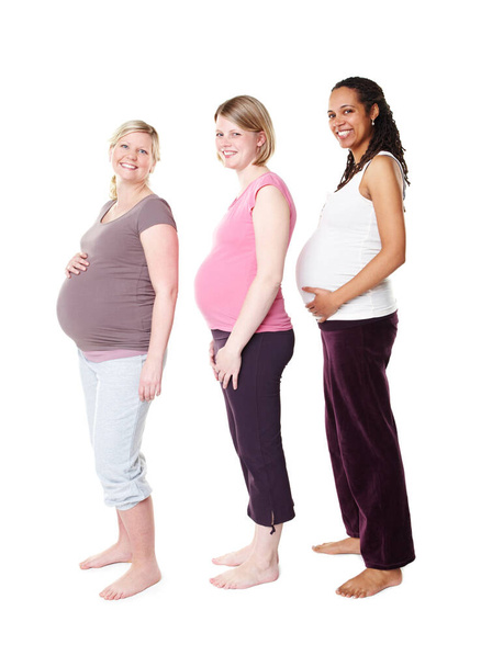 Comparing their baby bumps. Three pregnant women smiling broadly at the camera while standing against a white background holding their stomachs - Photo, Image