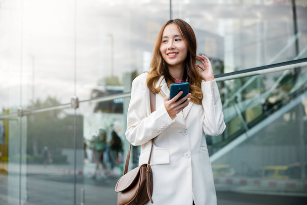 Retrato exitosa mujer de negocios sonriendo sosteniendo teléfono inteligente uso de chat aplicación en línea por la mañana, mujer de negocios asiática que trabaja con el teléfono móvil de pie contra el edificio de la calle frente a la oficina - Foto, imagen