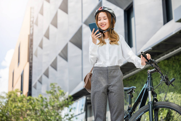 Asian smiling young woman with helmet hold mobile smart phone talk with business during go to office work at street with bicycle, Eco friendly, Lifestyle business female commuting outside in morning - Φωτογραφία, εικόνα