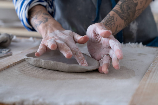 Closeup of working table in ceramics studio with female master with tattoo shaping wet clay plate. Woman potter sculpting decorating handmade bowl in workshop, cropped shot. Art and hobby concept - Foto, afbeelding