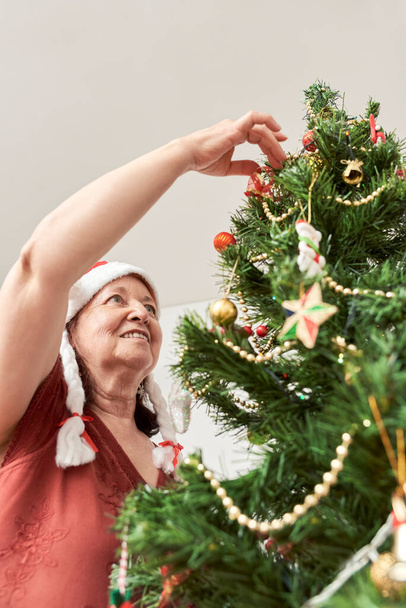 Szczęśliwa starsza Latynoska uśmiechnięta podczas dekoracji choinki w domu, w czerwonej czapce Świętego Mikołaja z białymi warkoczami. Radość świąt. - Zdjęcie, obraz