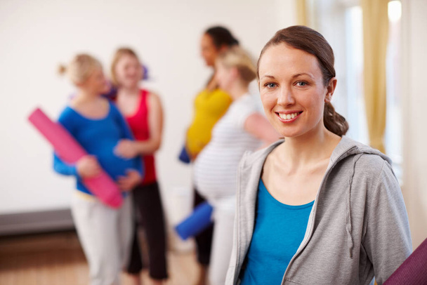 Relaxation exercise in perfect for pregnancy. A young pregnant woman standing in a gym holding an exercise mat with a group of women in the background - Photo, Image