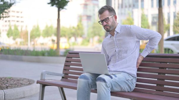 Дорослий чоловік з болем спини за допомогою ноутбука сидячи на відкритому повітрі на лавці
 - Фото, зображення