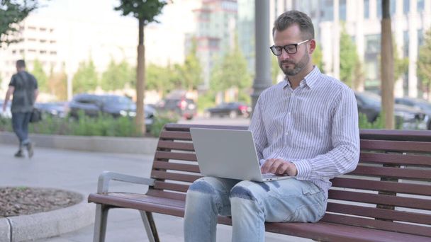 зайнятий дорослий чоловік за допомогою ноутбука сидить на відкритому повітрі на лавці
 - Фото, зображення