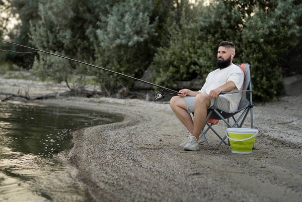 Hombre barbudo pescando en el lago sentado en embarcadero cerca del agua. concepto de ocio y personas - Foto, imagen