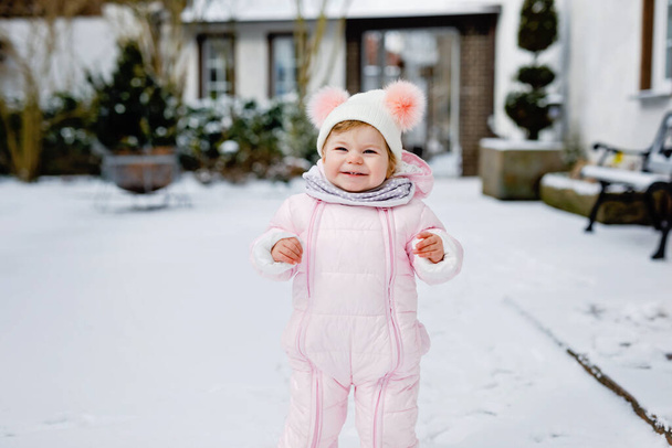Boldog kislány teszi az első lépéseket a szabadban télen a hóban. Aranyos kisgyerek, aki járni tanul. A gyerek jól érzi magát a hideg havas napokon. Meleg baba rózsaszín ruhát visel hóvédőt és bólogatós kalapot. - Fotó, kép