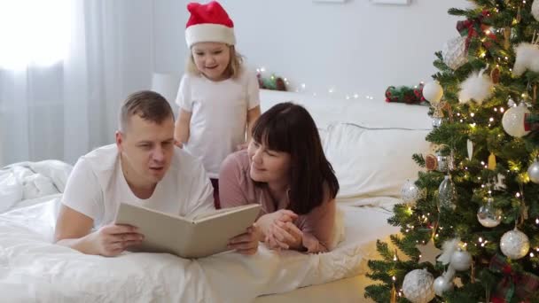 rodzina i koncepcja Bożego Narodzenia - młodzi rodzice czytający bajki małej córce w sypialni w pobliżu ozdobionej choinki - Materiał filmowy, wideo