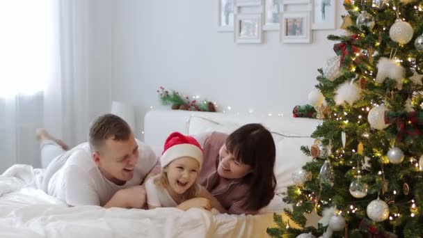 Familien- und Weihnachtskonzept - junge Eltern haben Spaß und kitzeln ihre kleine Tochter im Schlafzimmer neben geschmücktem Weihnachtsbaum - Filmmaterial, Video