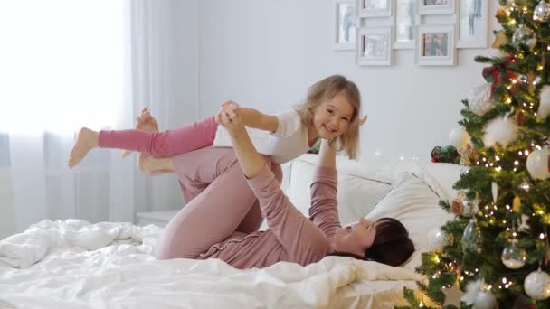 Aile ve Noel konsepti. Genç anne küçük kızıyla dekore edilmiş Noel ağacının yanındaki yatak odasında eğleniyor. - Video, Çekim