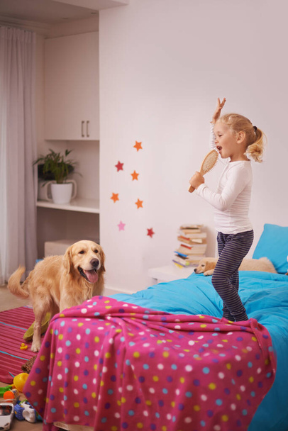 Hvězda ve výrobě. Malá holka skákání na posteli a zpěv do svého kartáče na vlasy, zatímco její pes se dívá - Fotografie, Obrázek