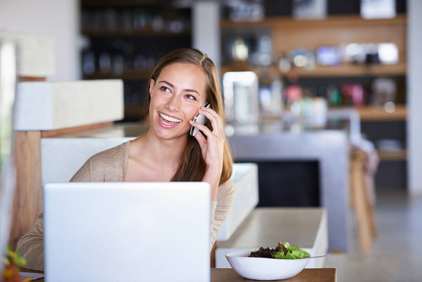 Социальная многозадачность. Счастливая молодая женщина разговаривает по телефону, сидя за столом дома - Фото, изображение